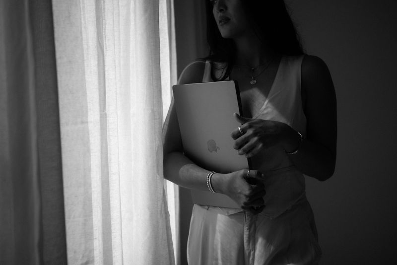 Black-and-white portrait of a woman by a sheer curtain, holding a laptop to her chest.