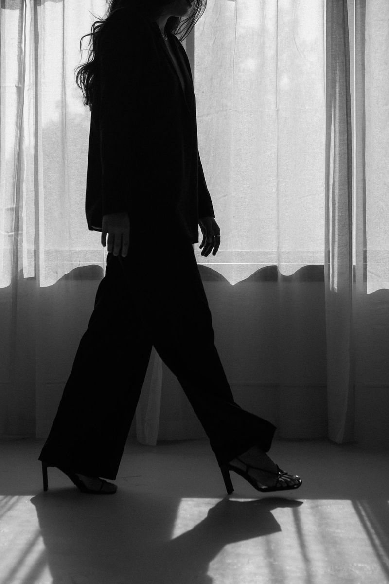 Black-and-white silhouette of a woman in a suit and heels walking past a sunlit curtain.