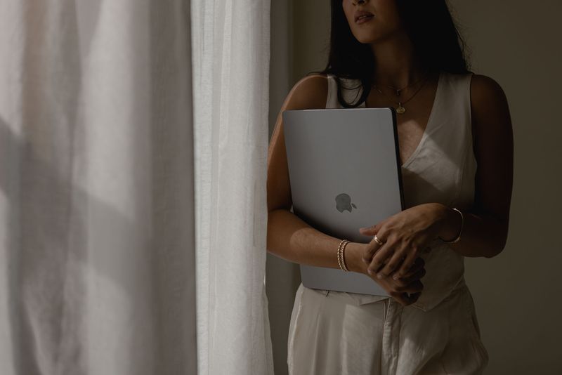 A woman in ivory linen holding a laptop beside a sunlit curtain, gold jewelry at her wrist.