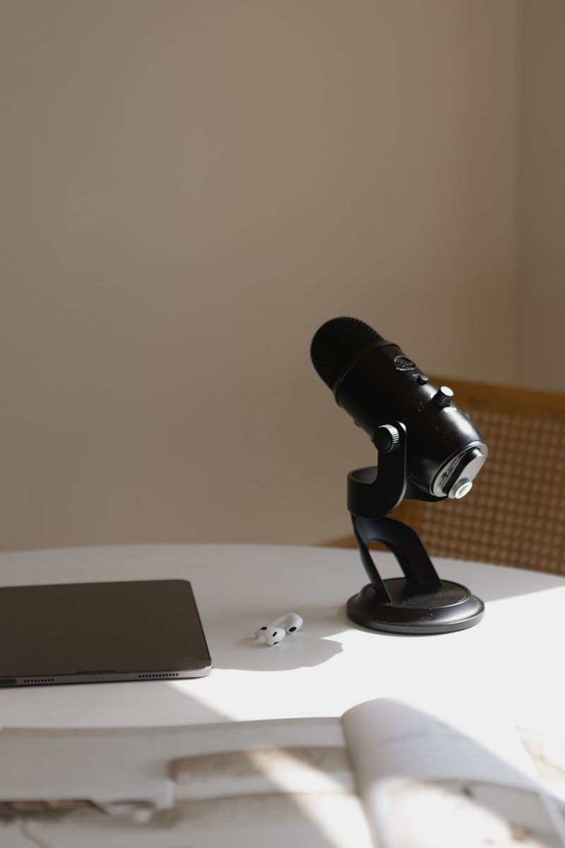 A black podcast microphone on a white table beside AirPods and a tablet.